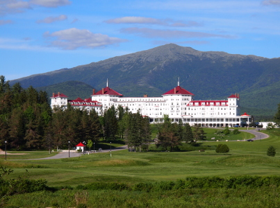 [The exterior of a four star resort. All white walls and a maroon roof set among evergreens with Mount Washington as a backdrop under blue skies with a few clouds. A small gatehouse and a large, well-manicured lawn sit in front of the resort.]